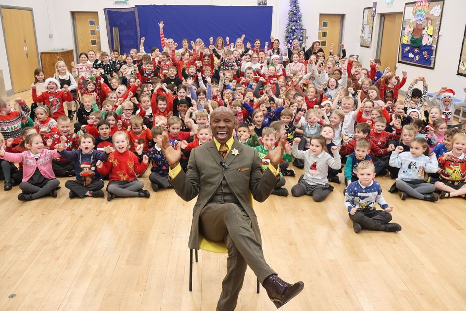 A man sits in a chair in front of a large group of children wearing Christmas sweaters.