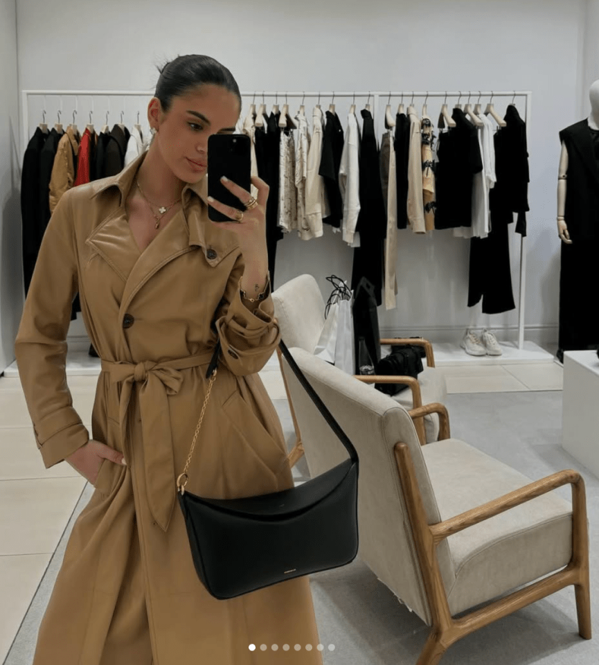 Woman in tan trench coat taking a selfie with black shoulder bag.