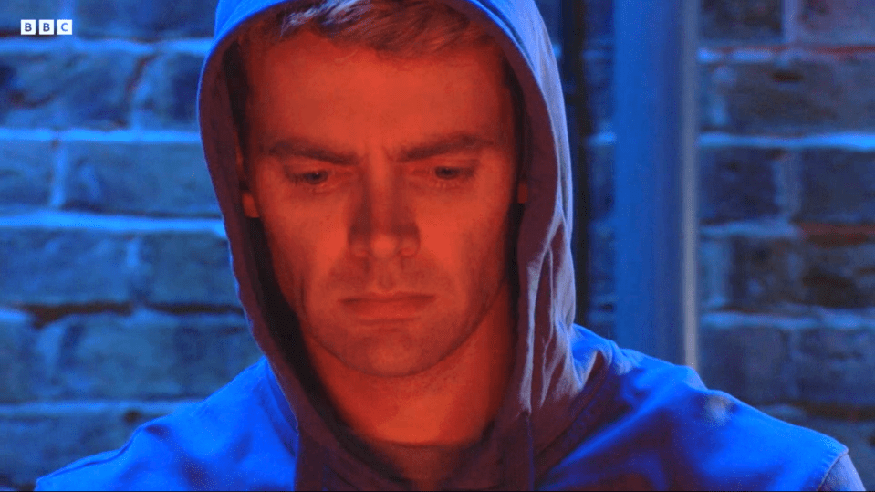 Screenshot of a man in a blue hoodie, his face illuminated in red light.