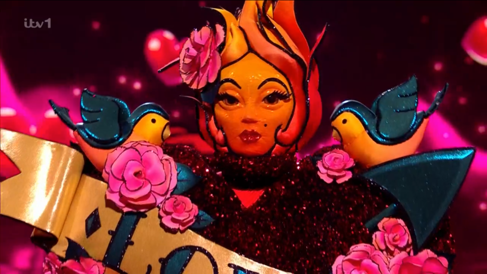 Masked singer contestant in a flame-haired costume with birds and roses.