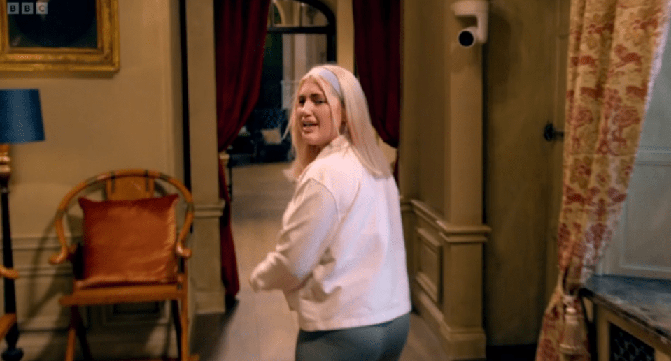 A blonde woman walking down a hallway in a large house.