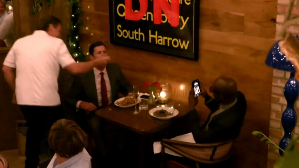 Screenshot of a man in a chef's uniform feeding another man at a restaurant table, while a third man takes a photo with his phone.