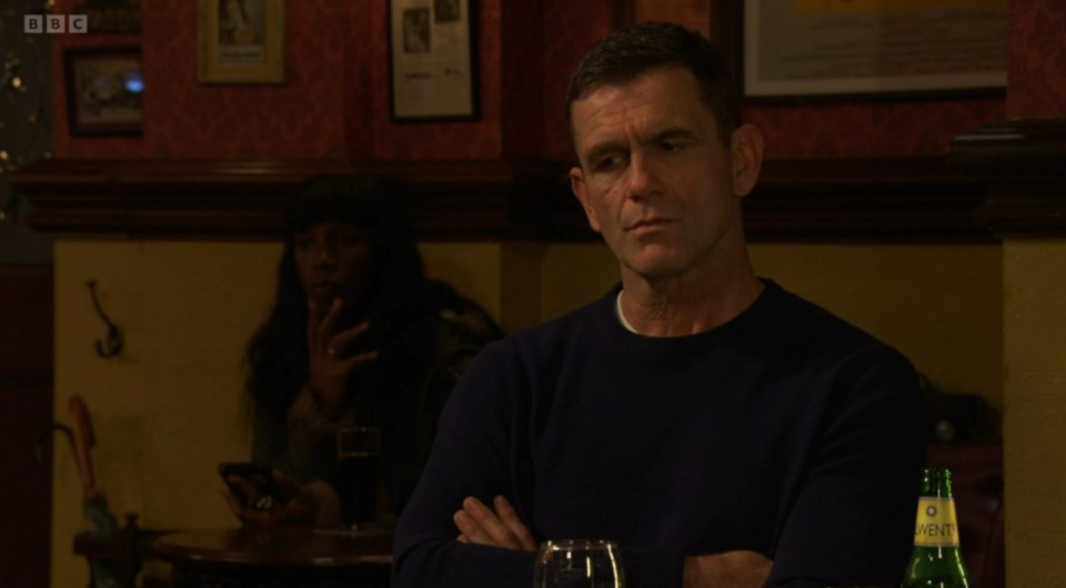 Screenshot of a man sitting in a pub with his arms crossed, a woman is visible in the background.