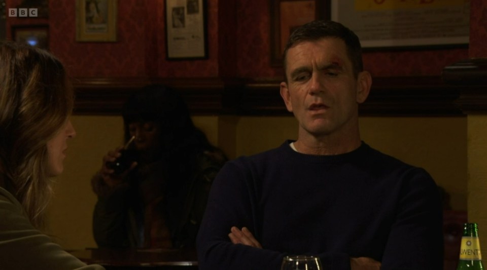 Screenshot of a man with a cut above his eye sitting in a pub, talking to a woman.