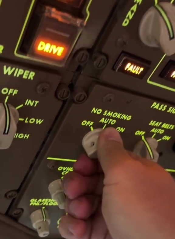 Close-up of an airplane cockpit's control panel; a hand adjusts a switch labeled "NO SMOKING AUTO."