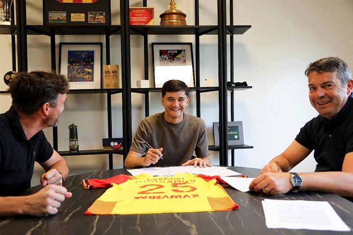 A young man signing a contract, flanked by two other men.  A soccer jersey lies on the table.