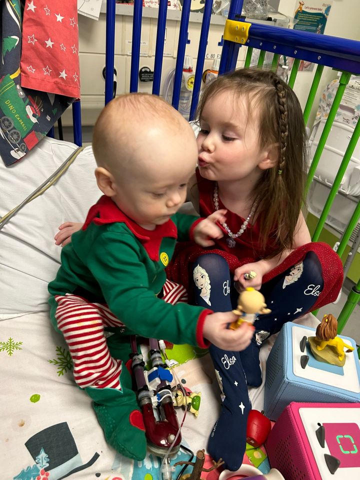 A young girl kisses her baby brother, who is connected to a Berlin Heart machine.