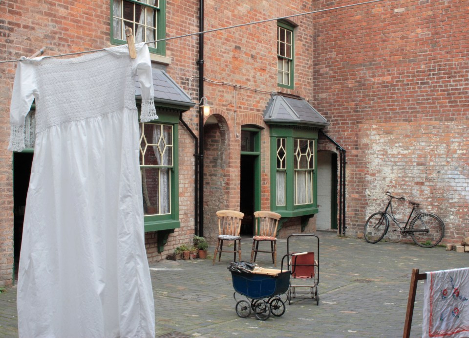 A white dress hangs on a clothesline in a brick courtyard with chairs, a baby carriage, and a bicycle.