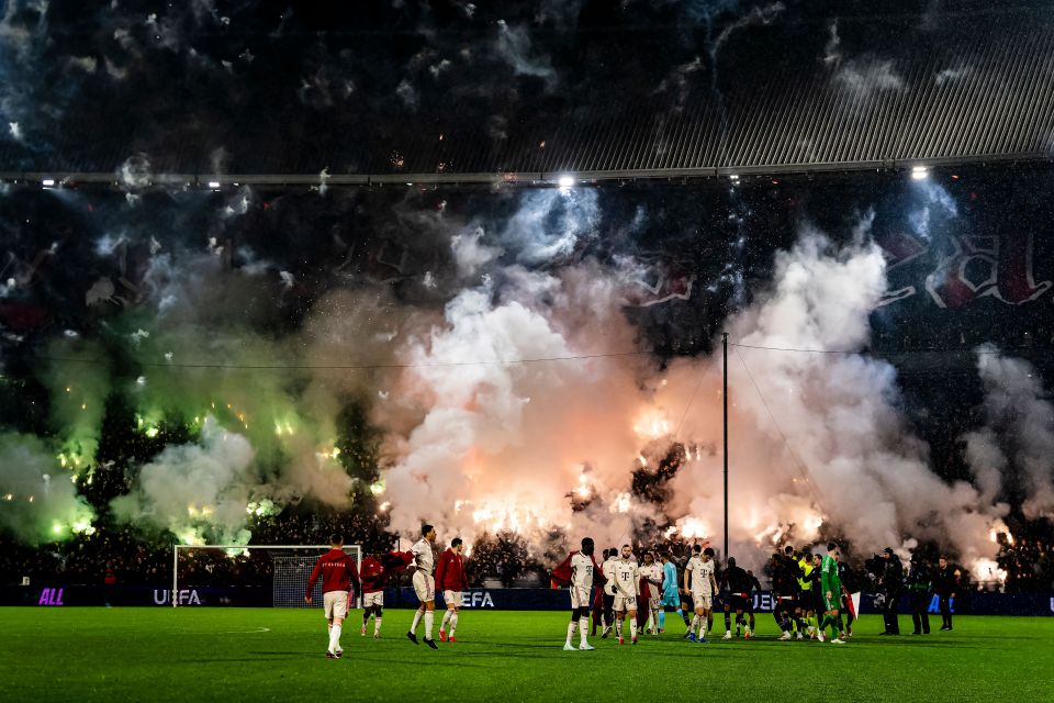 Soccer players walk on the field during a game, surrounded by smoke and flares from the crowd.