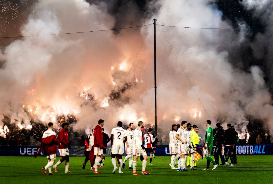 Soccer players on the field during a game with a large amount of smoke and flares from the crowd in the background.