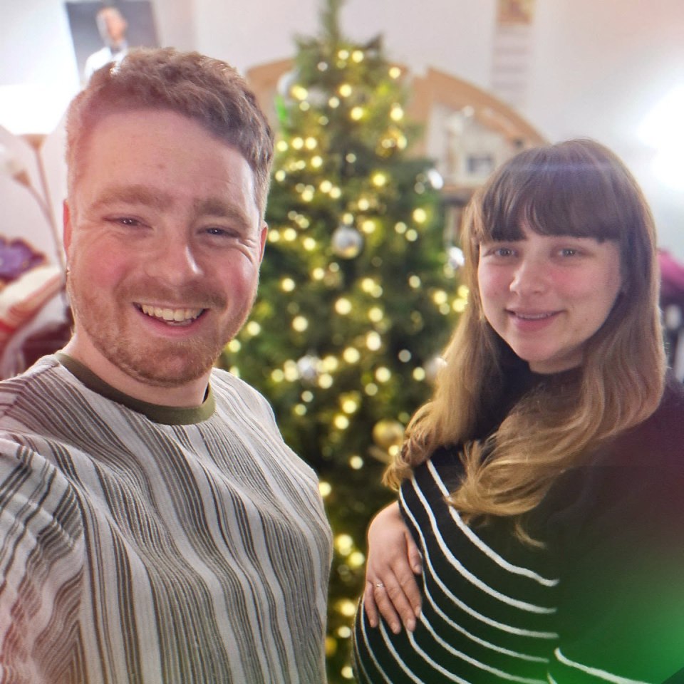 Tom Ball and his wife, pregnant, in front of a Christmas tree.