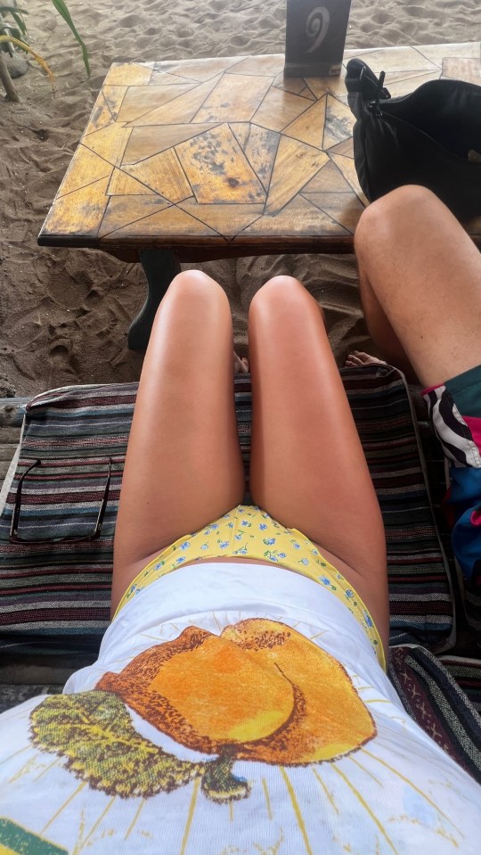 Woman in bikini bottom and peach-print shirt sitting at beachside table.