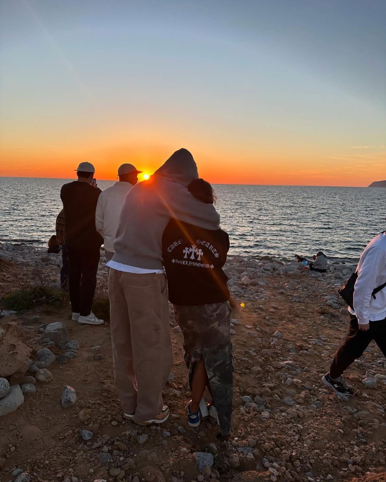 Ruben Dias and Maya Jama embracing at sunset by the ocean.