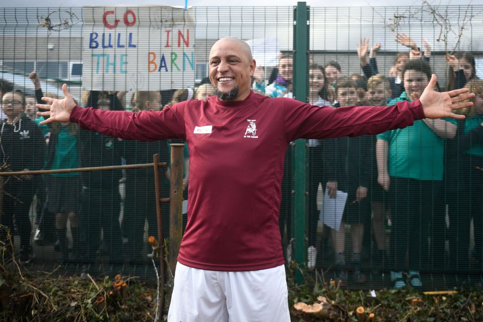 Roberto Carlos, former Real Madrid player, smiles and poses with arms outstretched in front of children.