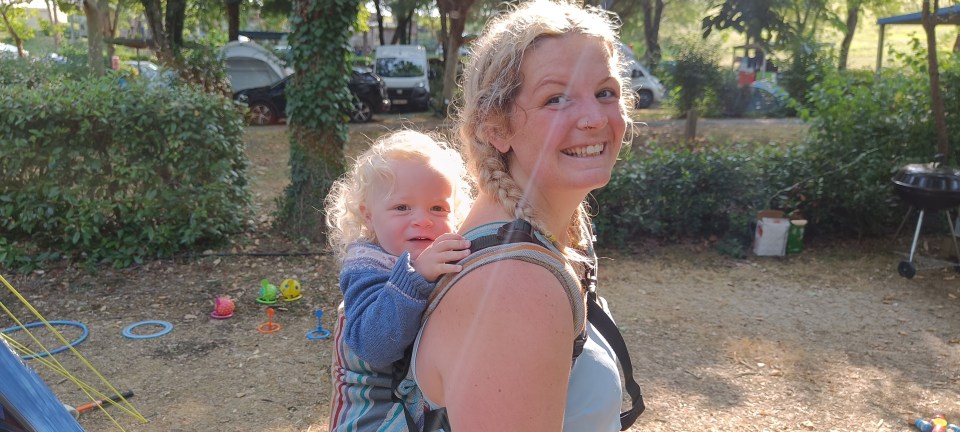 Woman carrying toddler in a baby carrier at a campsite.