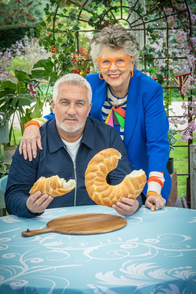 Paul Hollywood and Prue Leith, judges from The Great British Bake Off, holding a large braided loaf of bread.