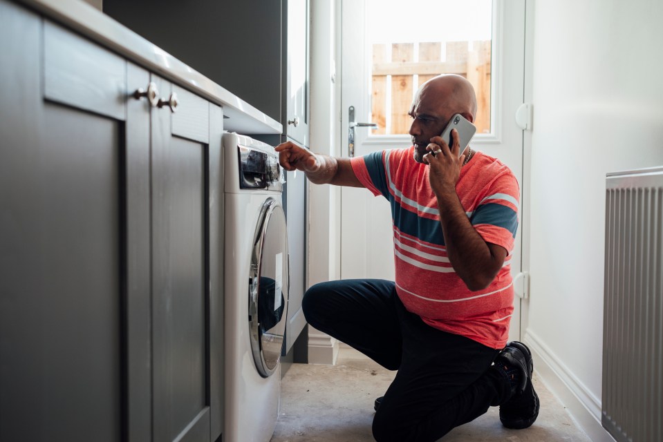 A man on a phone calls for appliance repair while kneeling next to a broken washing machine.