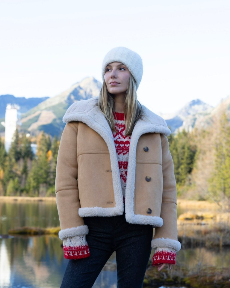 Woman in a sheepskin coat and knit hat by a lake in the mountains.