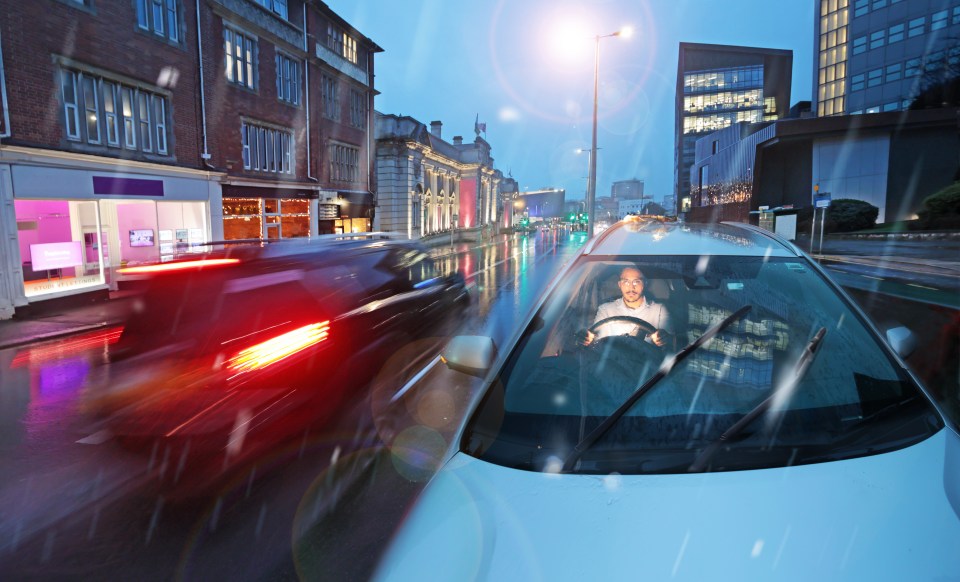 A man driving an electric car through a city in the rain at dusk.