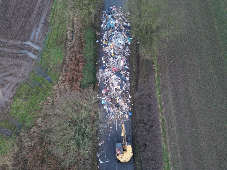 Aerial view of a construction vehicle removing a large pile of illegally dumped waste from a road.