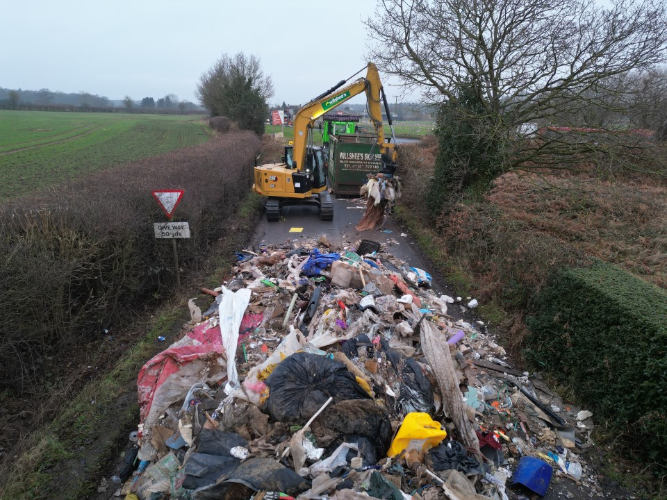 Excavator removing a large pile of illegally dumped waste from a road.