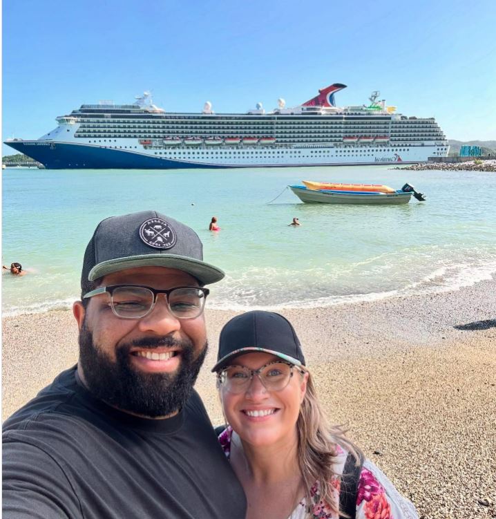 Couple taking a selfie on a beach with a cruise ship in the background.