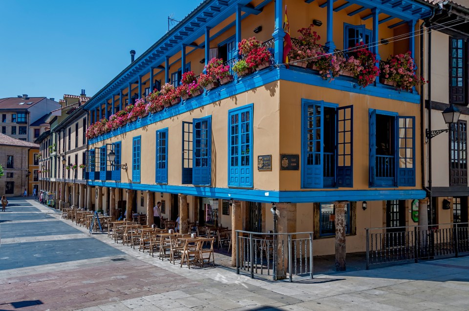 A yellow building with blue balconies and windows, flower boxes, and outdoor seating.