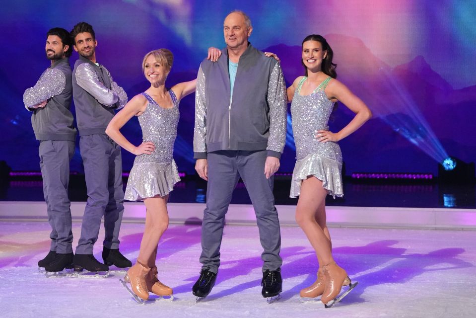 Five people on ice skates on a rink, two women in sparkly dresses, two men in gray jackets, and a man in a gray jacket in the center.