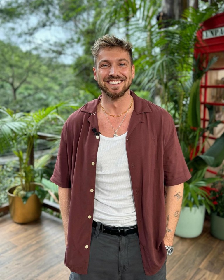 Man smiling in maroon shirt, standing outdoors near a red telephone booth.