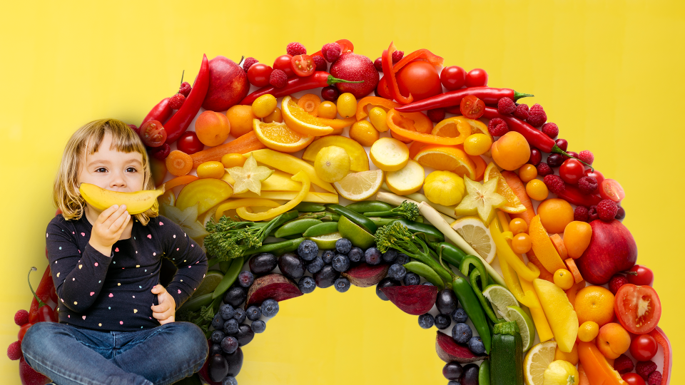 Child holding a banana in front of a rainbow of fruits and vegetables.