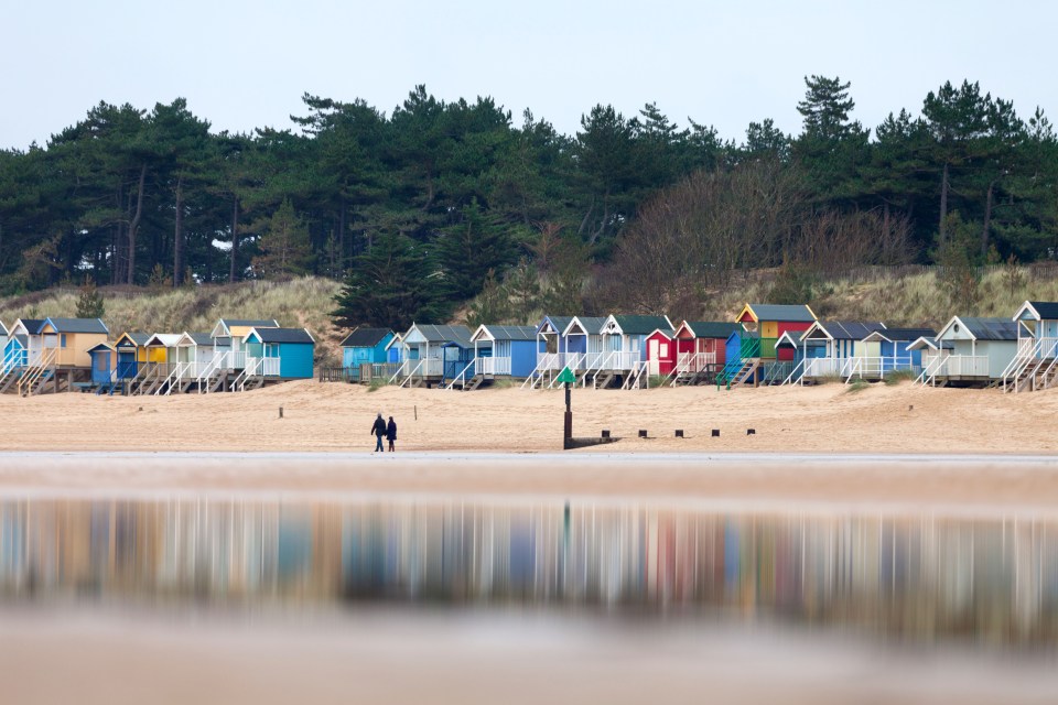 Couple walking on beach with colorful beach huts and their reflection in the water.