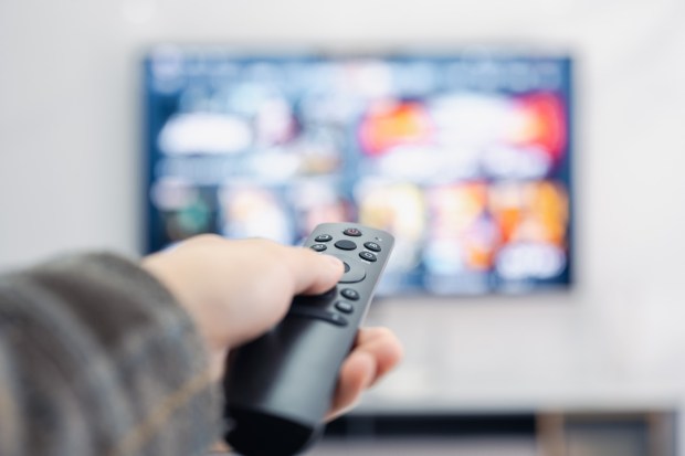 Hand holding a TV remote control in front of a blurred television screen.