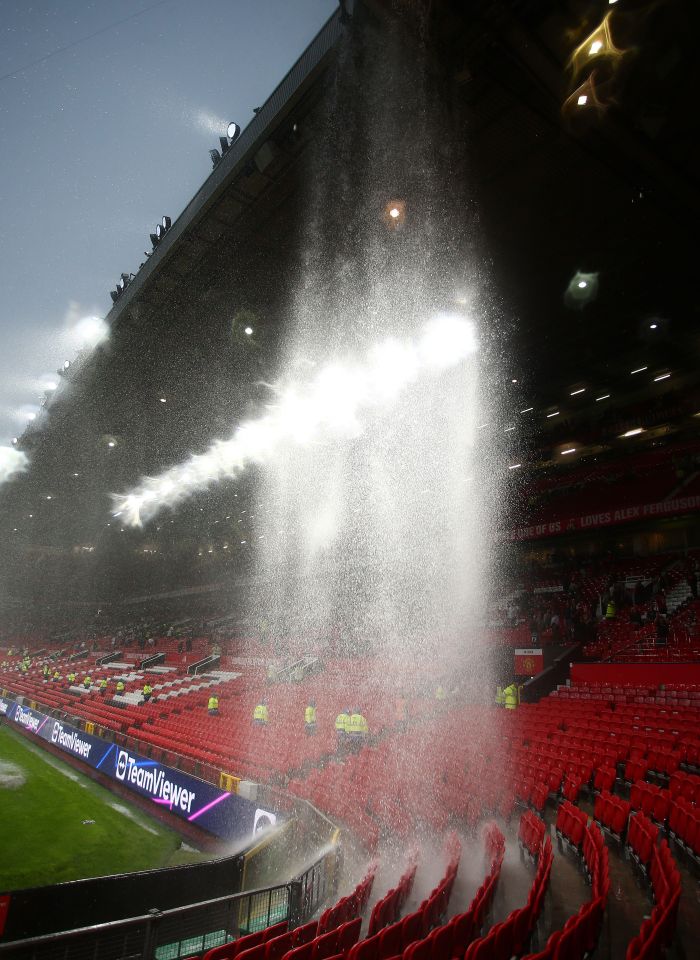 Water spraying from the roof of Old Trafford stadium onto empty red seats.