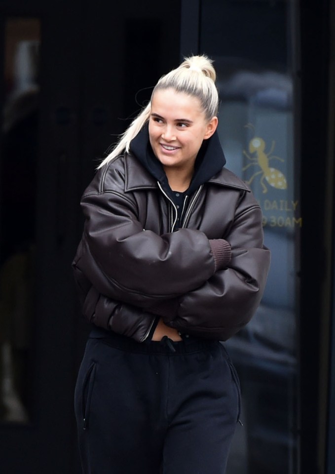 Molly Mae smiling while wearing a brown leather jacket and black sweatpants.