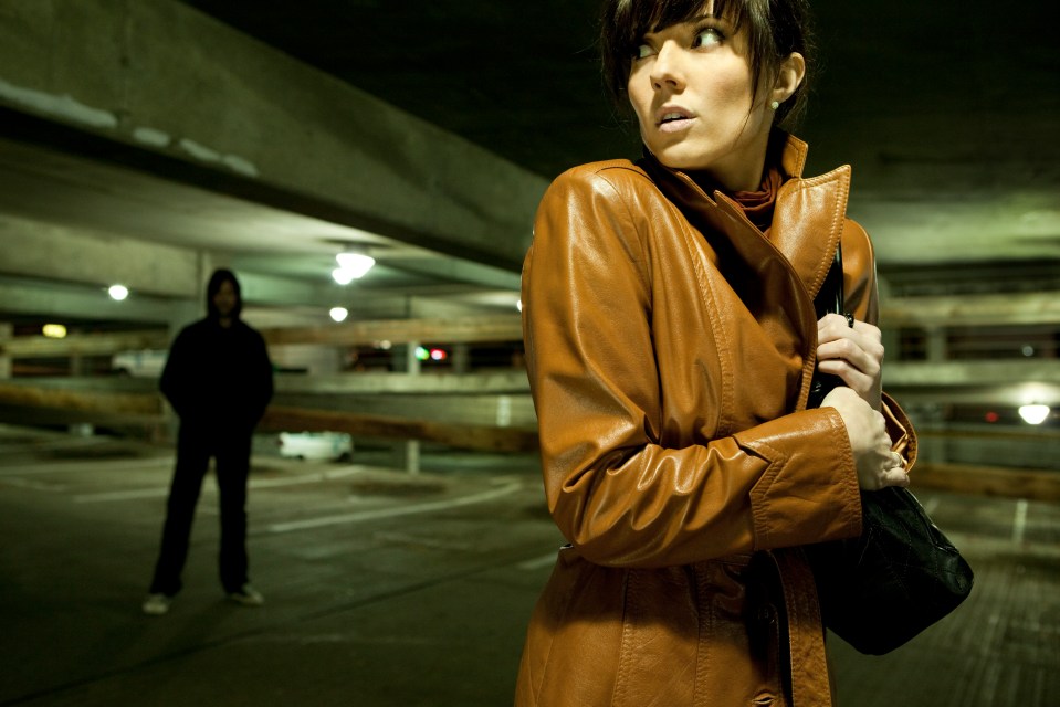 A fearful woman clutches her purse in a dimly lit parking garage, looking over her shoulder at a man in the background.