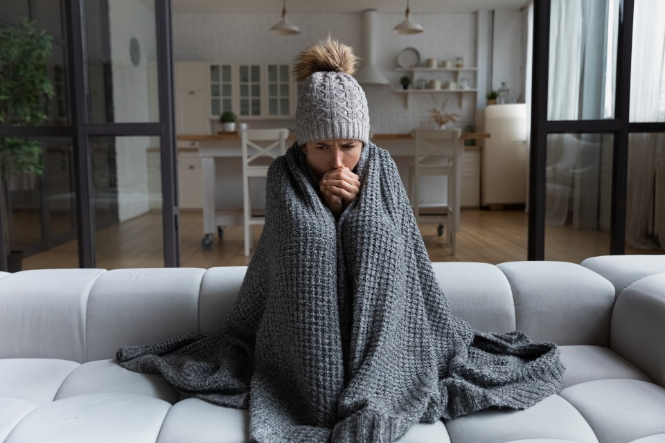 A woman sits on a couch wrapped in a blanket, shivering from the cold.