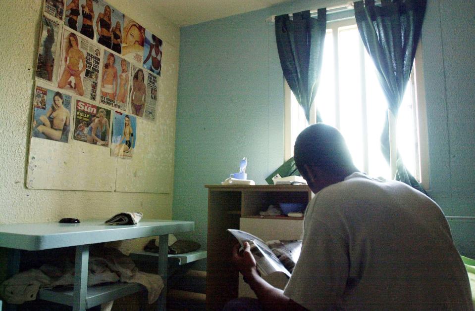 A young man sits in a prison cell reading a magazine; a wall is covered with tabloid clippings.