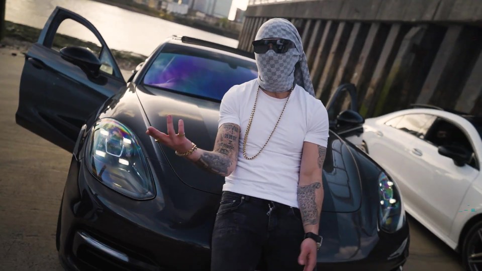 Man in a balaclava standing between two luxury cars.