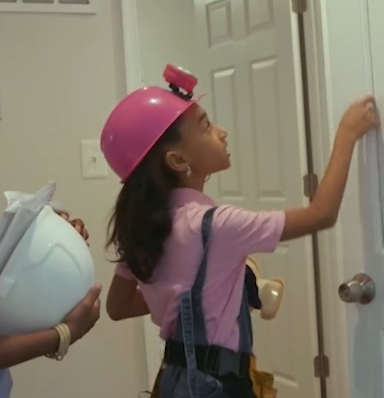 A young girl wearing a pink hard hat inspects a door frame.