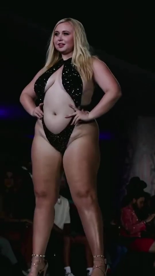 Plus-size model walking a runway in a sparkly black swimsuit.