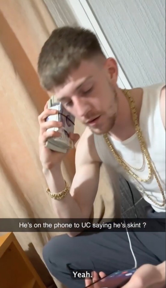 A man wearing gold chains holds a stack of cash to his ear while speaking on the phone, claiming to be struggling financially.