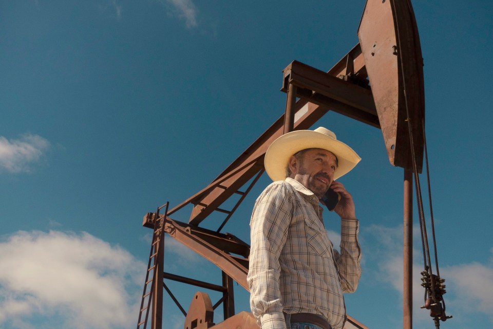 Billy Bob Thornton as Tommy Norris from the series "Landman," on a cell phone, standing near an oil derrick.