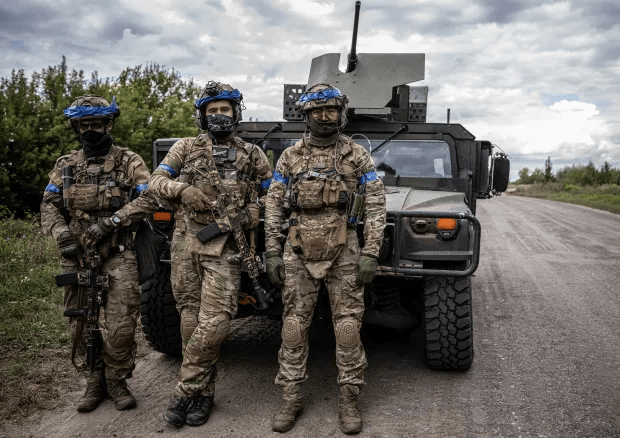 Three soldiers in camouflage uniforms stand with rifles near a military vehicle.