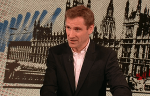 Man in suit speaking in front of a graphic of the Houses of Parliament.