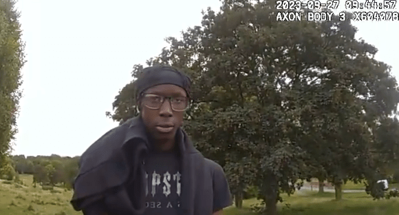 A young man wearing glasses and a black bandana stands outdoors.