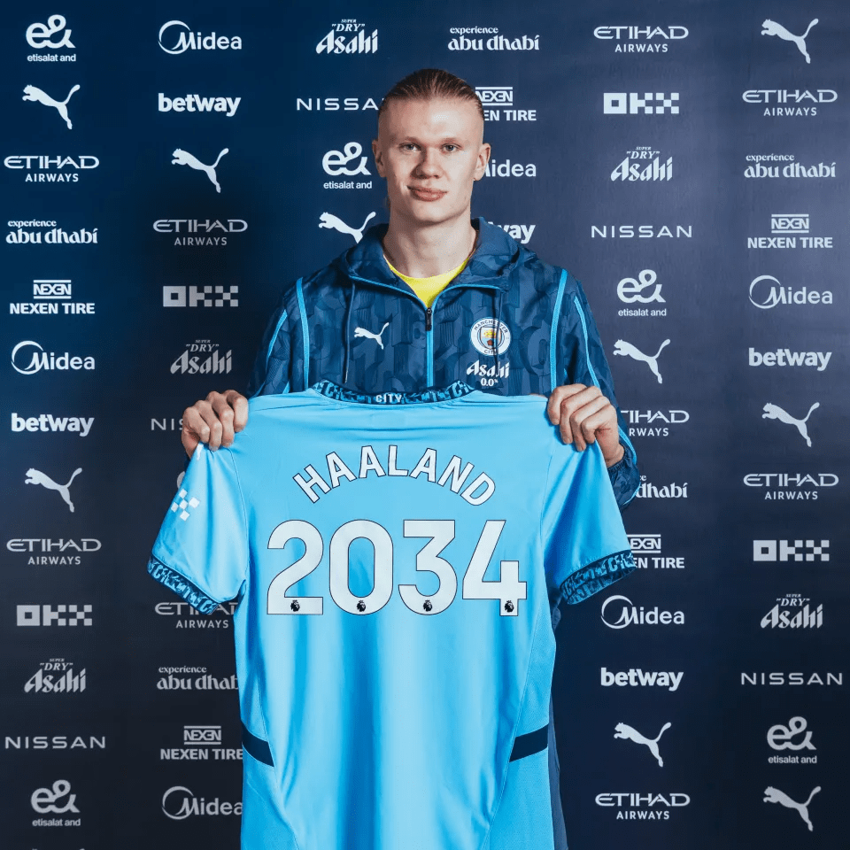 Erling Haaland holding a Manchester City jersey with "Haaland 2034" on the back.