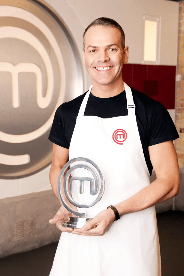 A smiling chef in an apron holds a MasterChef trophy.