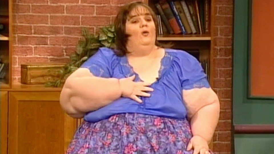 A morbidly obese woman in a blue shirt and floral skirt speaks on a talk show set.