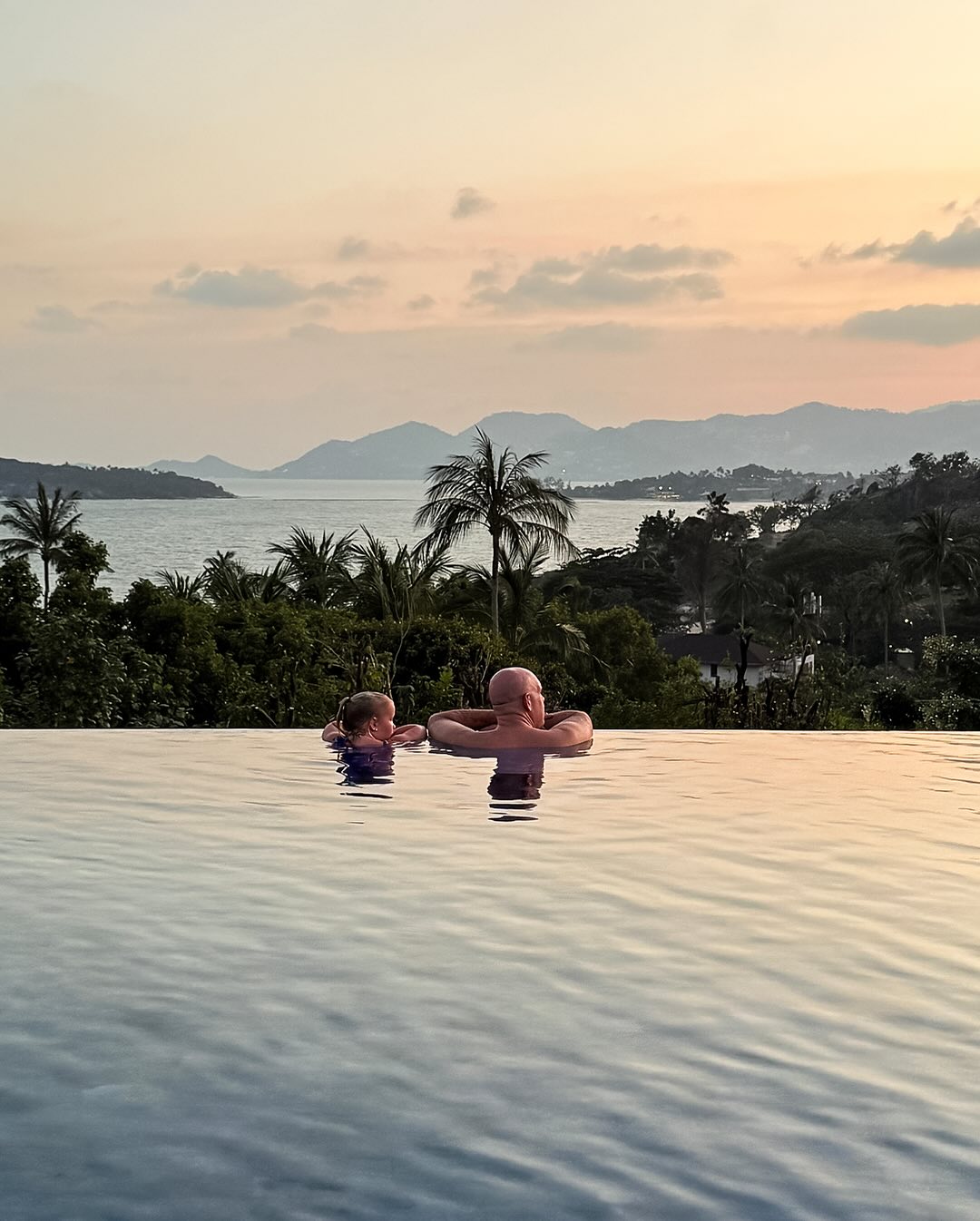 A man and child in an infinity pool overlooking the ocean at sunset.