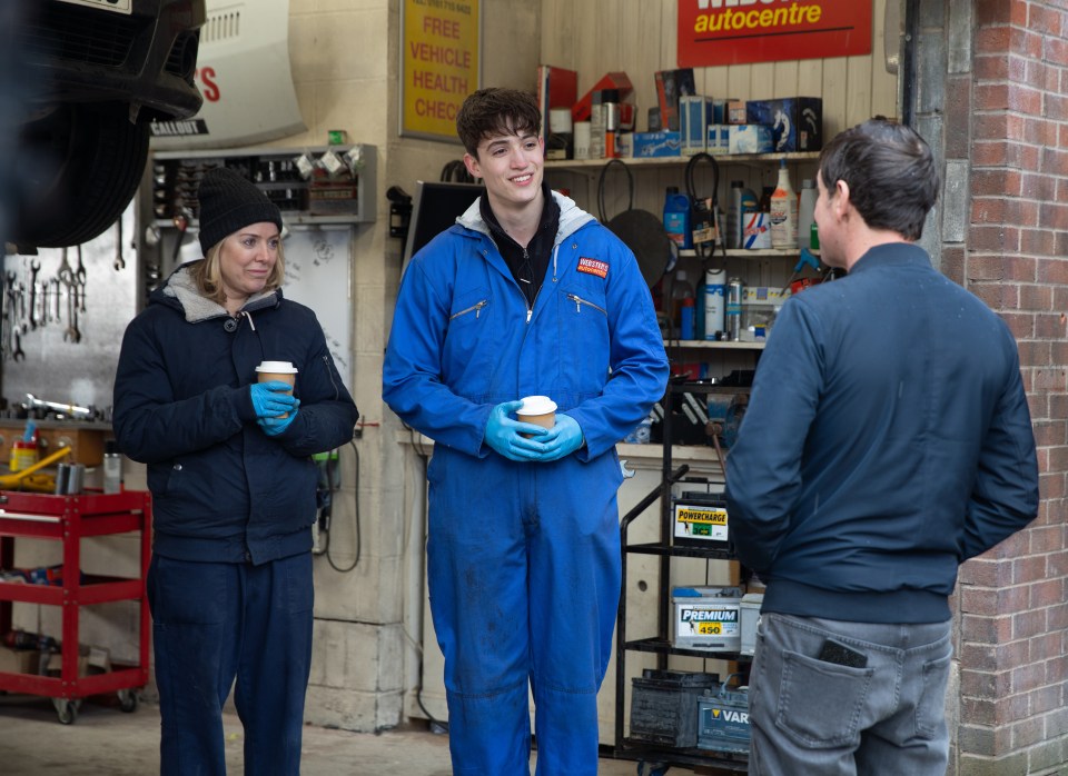 Three people, two women and a man, stand in a garage and talk while holding coffee cups.